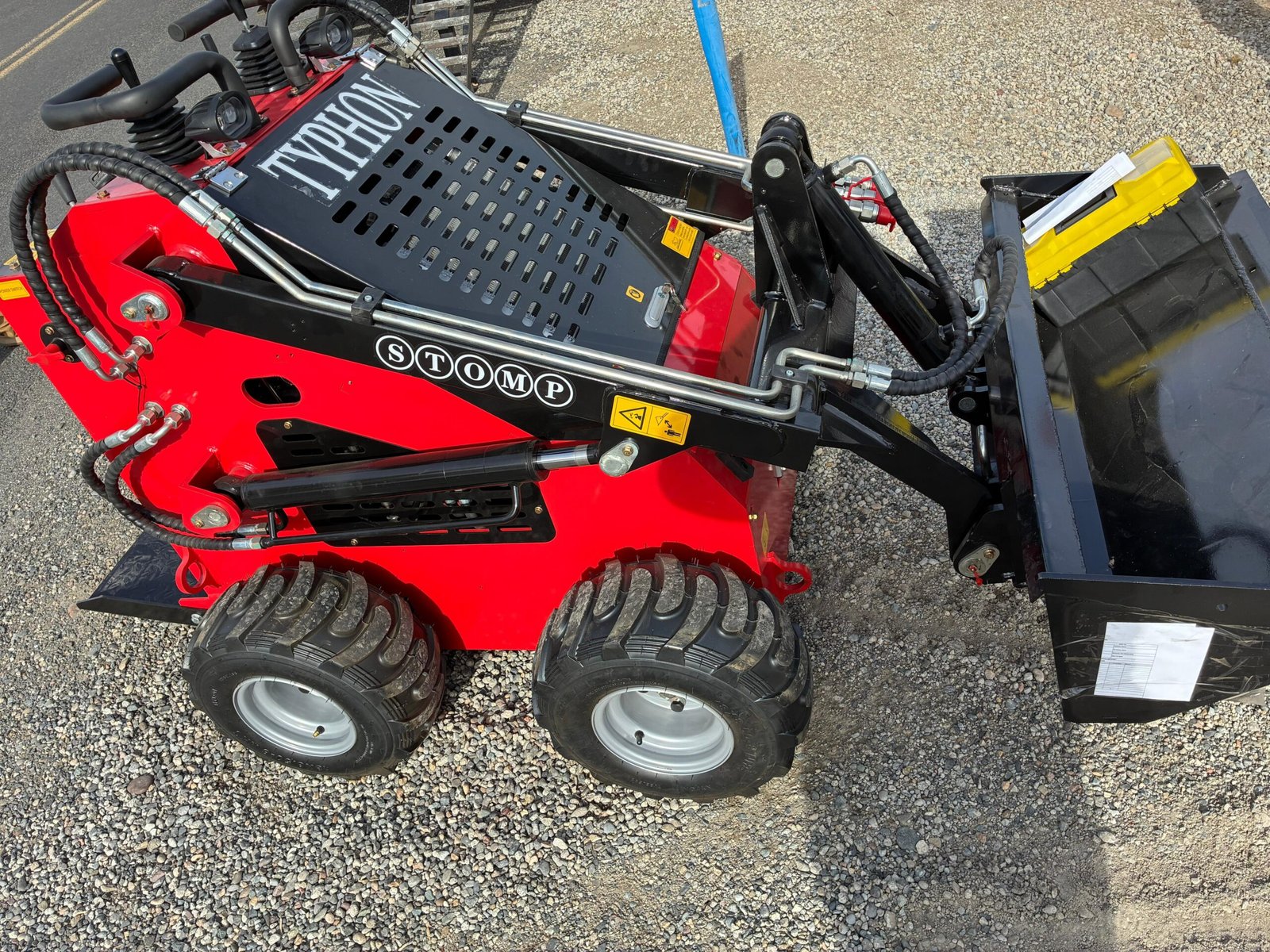 Close-up of red and black heavy machinery with hydraulic hoses, a perforated guard, and four rugged tires on a gravel surface.