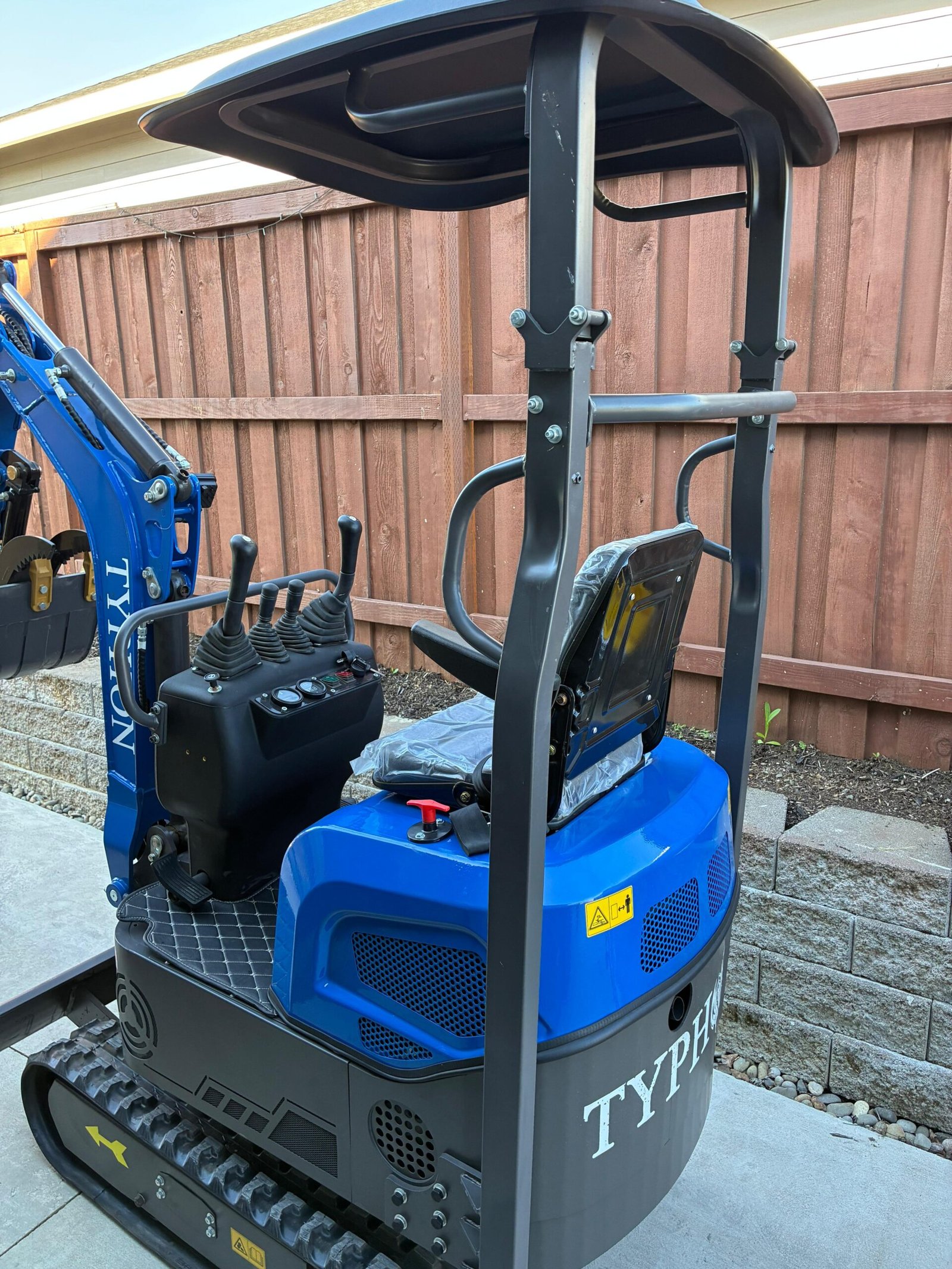 Blue mini excavator with black seat and control levers, parked beside a wooden fence in a backyard setting.
