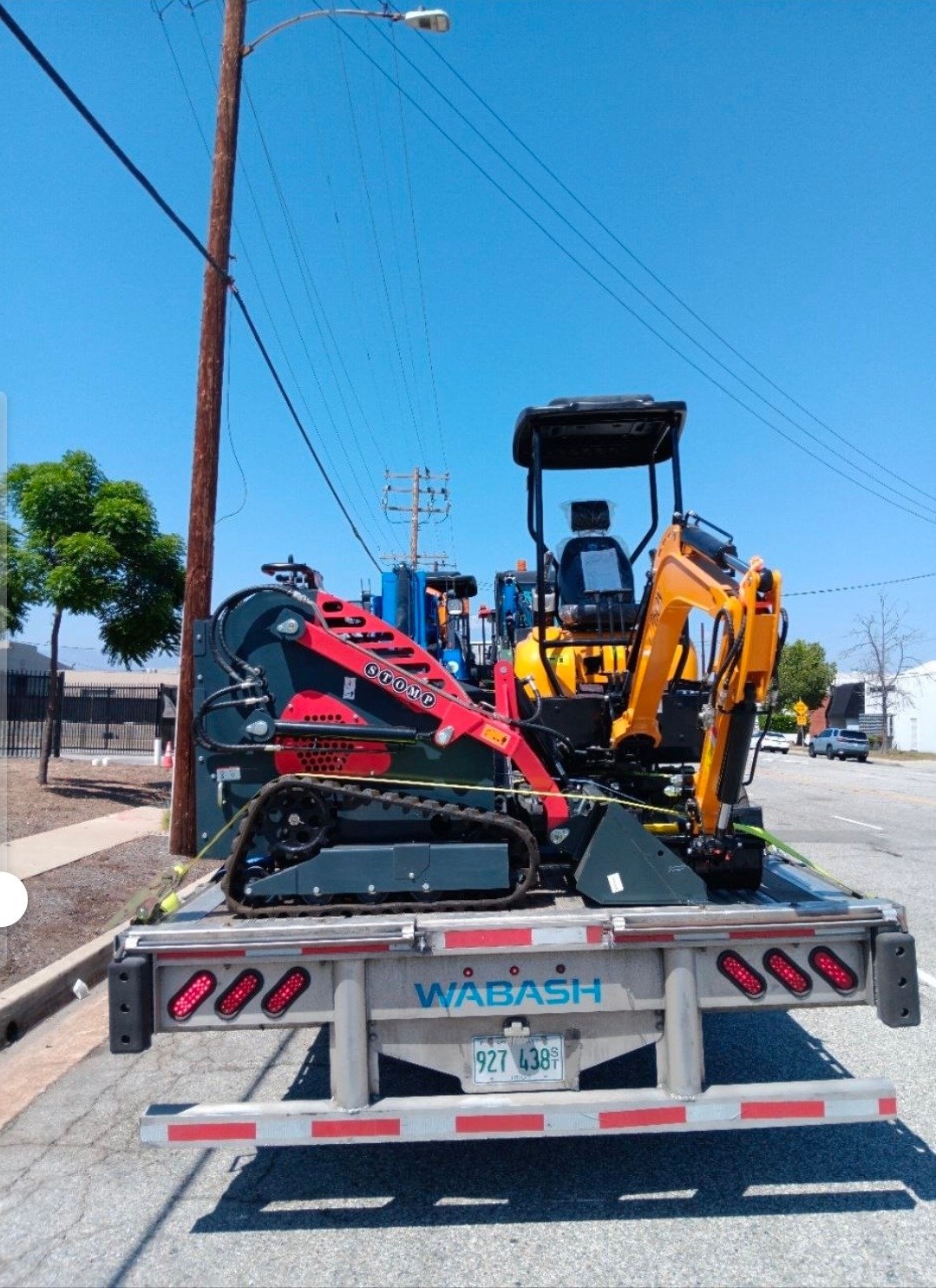 Trailer loaded with yellow mini excavator and red stump grinder on a sunny street.